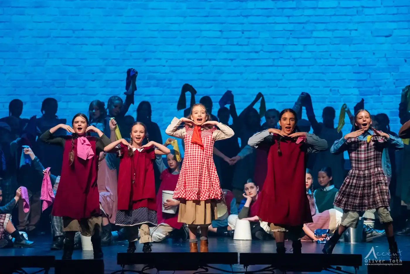 Young Pelican performers singing and dancing against a glowing blue brick wall